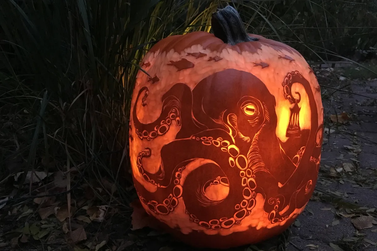 A pumpkin in partial darkness, lit up from within to show a detailed carving of a large octopus holding a lantern