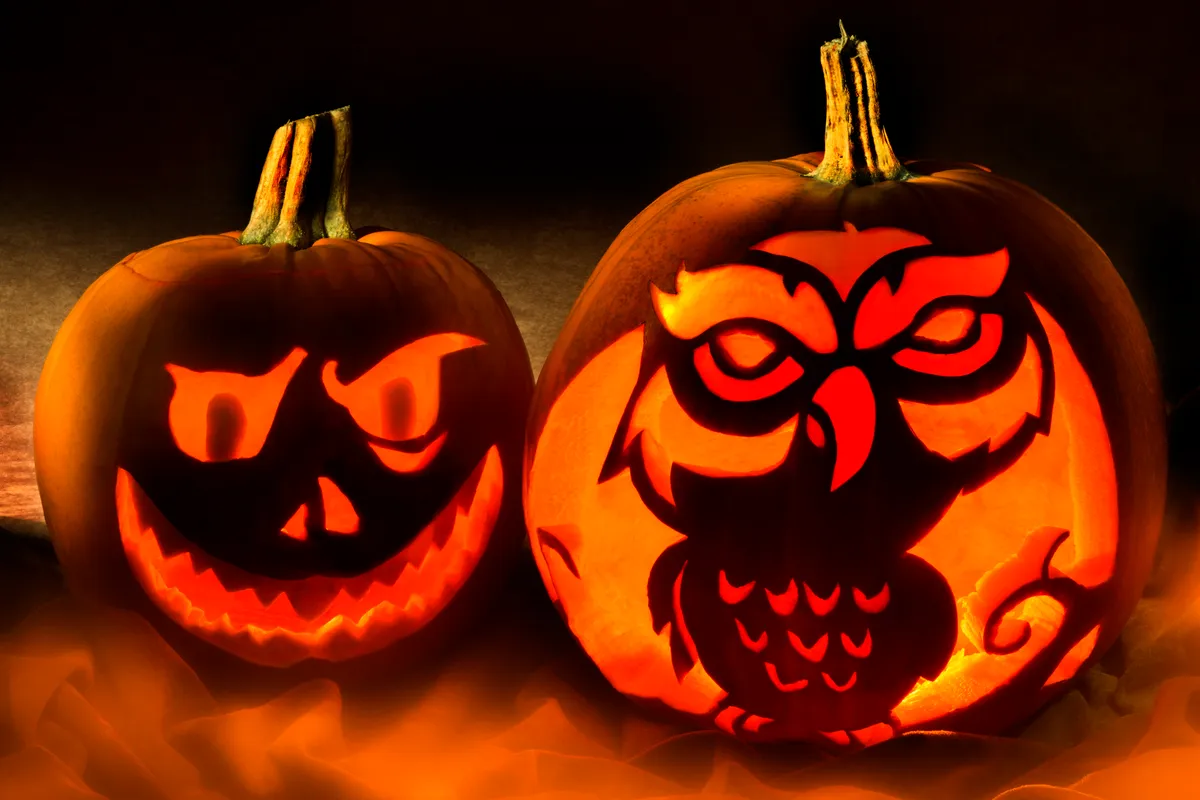 Two pumpkins in semi-darkness, lit up from within to show an owl carving on one pumpkin and a smiling spooky face on the other.