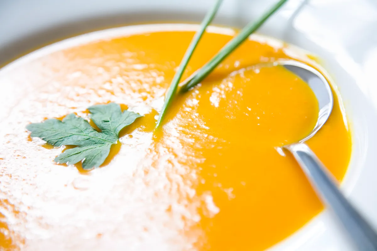 Creamy pumpkin soup in a bowl, with a silver spoon in it and a herbs on top.