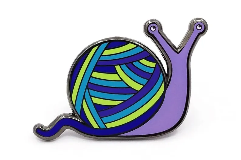 A snail enamel pin, with a purple body and a blue-green shell that looks like yarn, against a white background.
