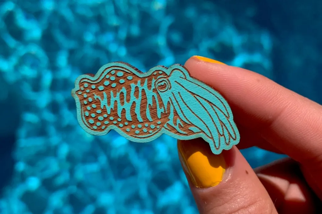 A blue wooden pin badge of a cuttlefish, with indents showing the natural wooden colour beneath, held above the blue waters of a swimming pool by a hand with yellow-orange painted fingernails.