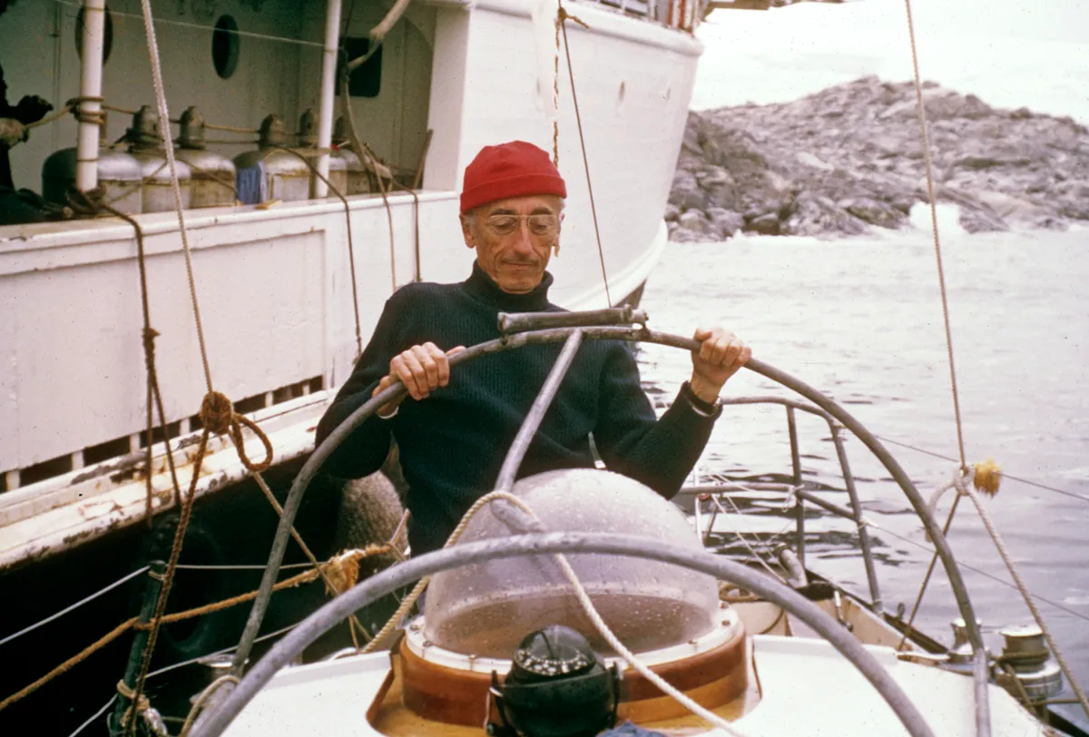 Jacques Cousteau, wearing a dark turtleneck jumper and his red hat, standing on a small boat which is next to a larger boat (likely the Calypso), with water and rocks in the background.