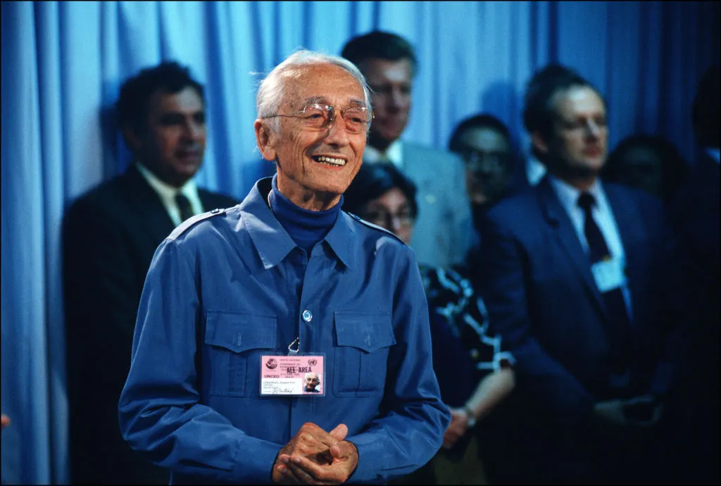 Jacques Cousteau (in his 80s) wearing a blue shirt over a blue turtleneck indoors at a conference, standing in front of a number of other delegates.
