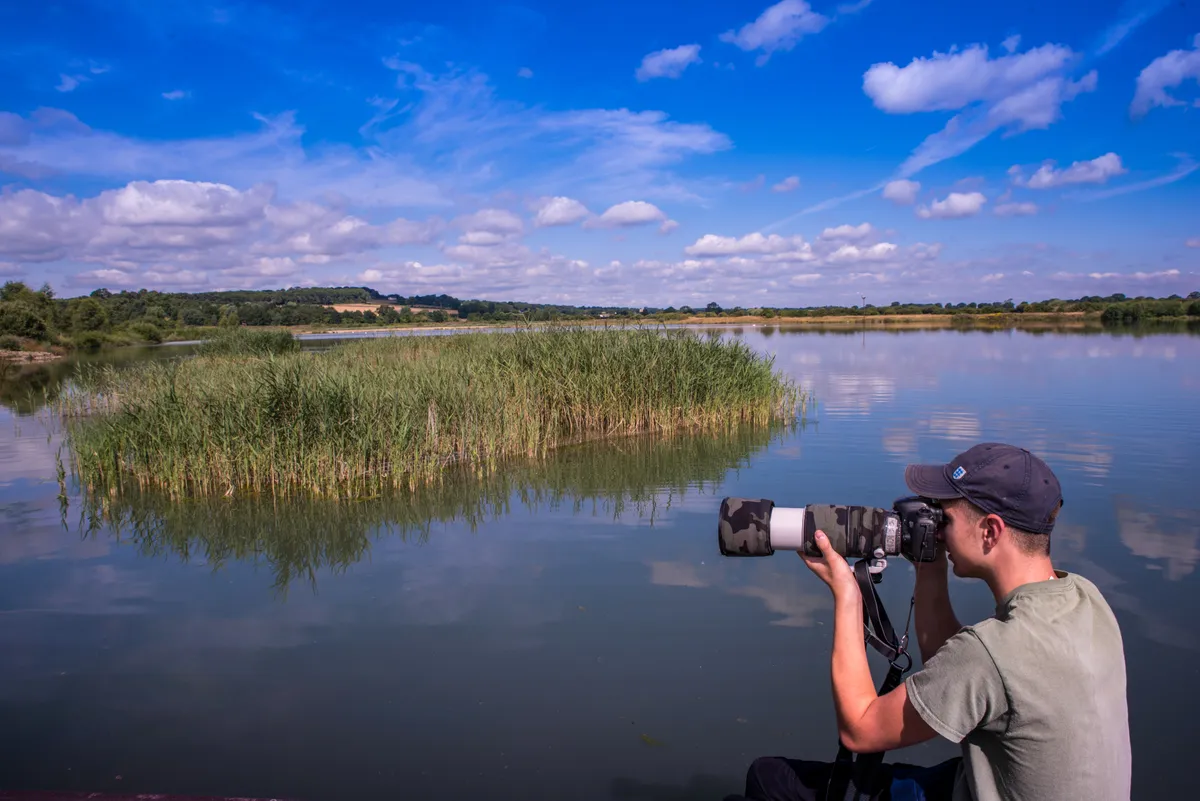 A boy in his mid to late teens crouches in front of a lake, hold a camera up to his eye to take photos