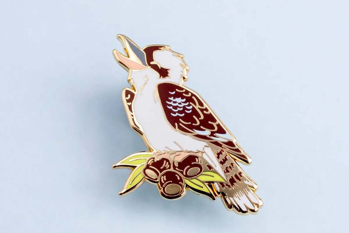 A kookaburra enamel pin badge will gold inlay, design of a kookaburra on a branch calling, against a white background.