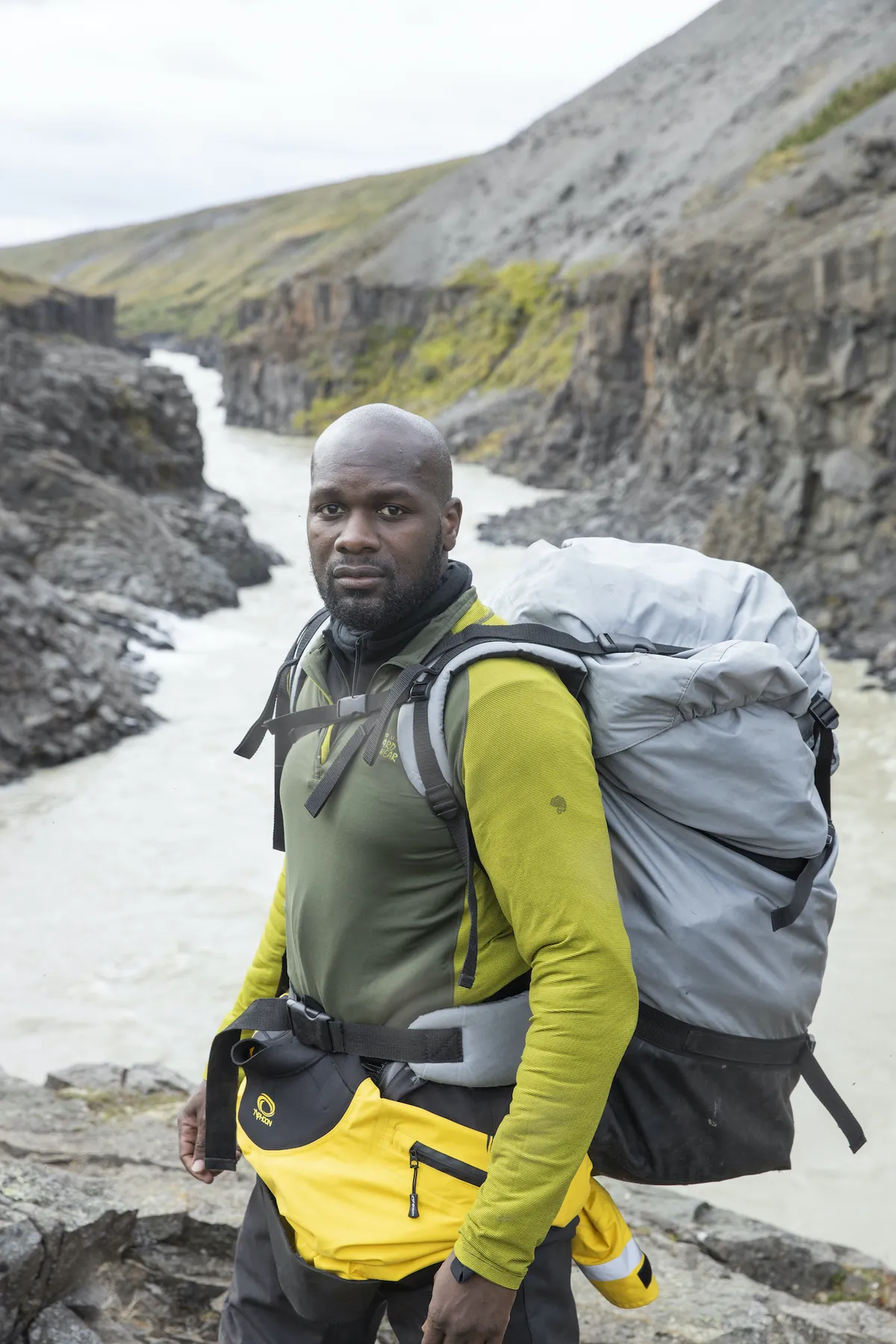 Dwayne Fields looks into the camera, wearing outdoors clothing and a very large rucksack. Behind him, a river cuts through a gorge.