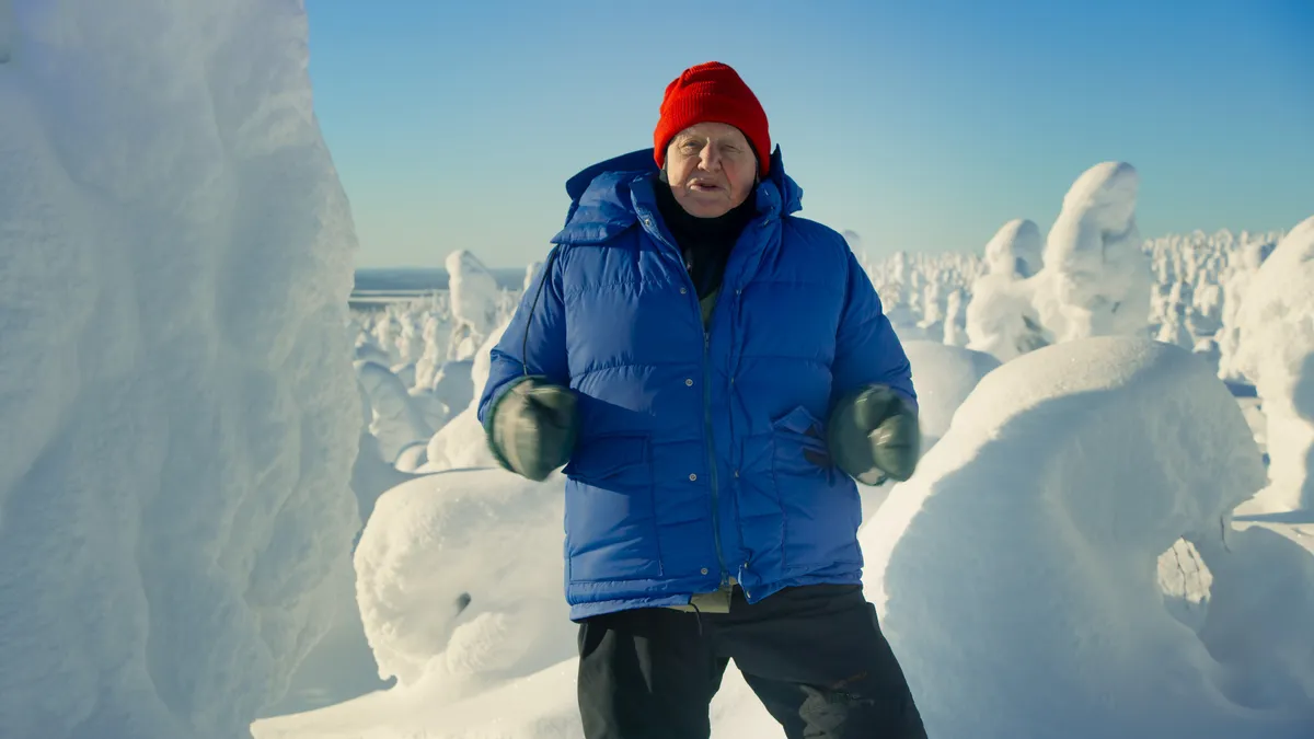 Sir David Attenborough facing the camera, wearing lots of layers and standing in front of snow-covered scene.