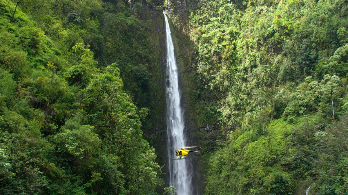 A yellow helicopter flying between trees in front of a waterfall.