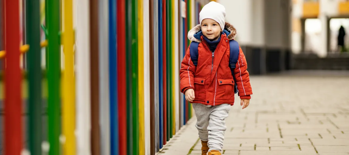 2024-kindergartenweg-wann-kann-kind-alleine-in-den-kindergarten-fritz-und-fraenzi-evelin-hartmann Ein kleine Junge mit roter Jacke und weisser Mütze auf dem Weg in den Kindergarten.
