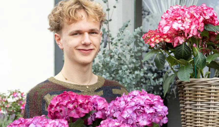 berufswahl-dominic-koch-lehre-florist-ich-will-kunden-gluecklich-machen-fritz-und-fraenzi Ein junger Mann steht vor einem Blumenladen wo er als Florist seine Lehre absolviert. Er lächelt leicht.
