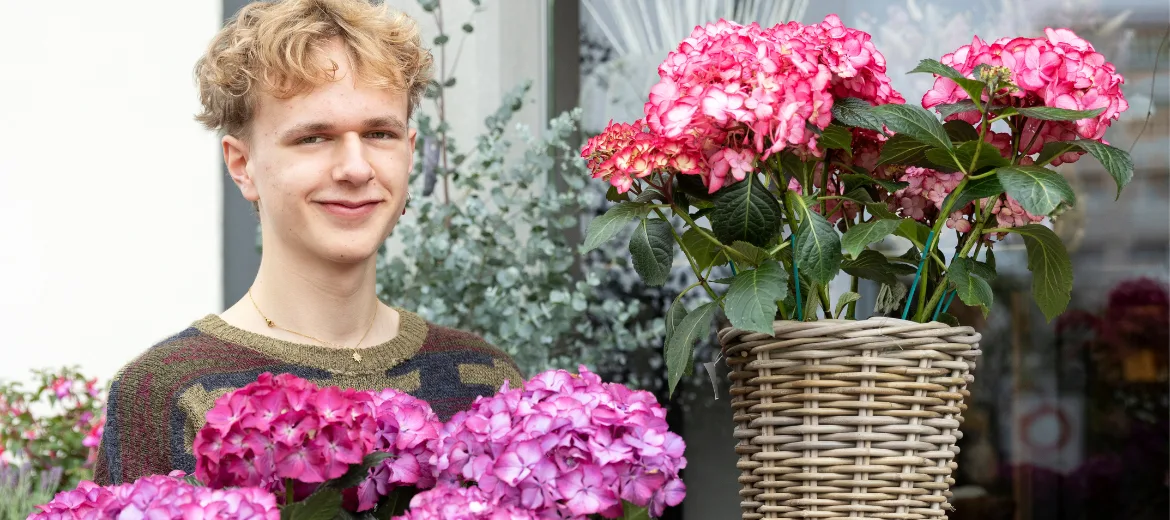 choix-de-profession-dominic-cuisinier-apprentissage-floriste-je-veux-faire-des-clients-heureux-fritz-et-fraenzi Un jeune homme se tient devant un magasin de fleurs où il fait son apprentissage de fleuriste. Il sourit légèrement.