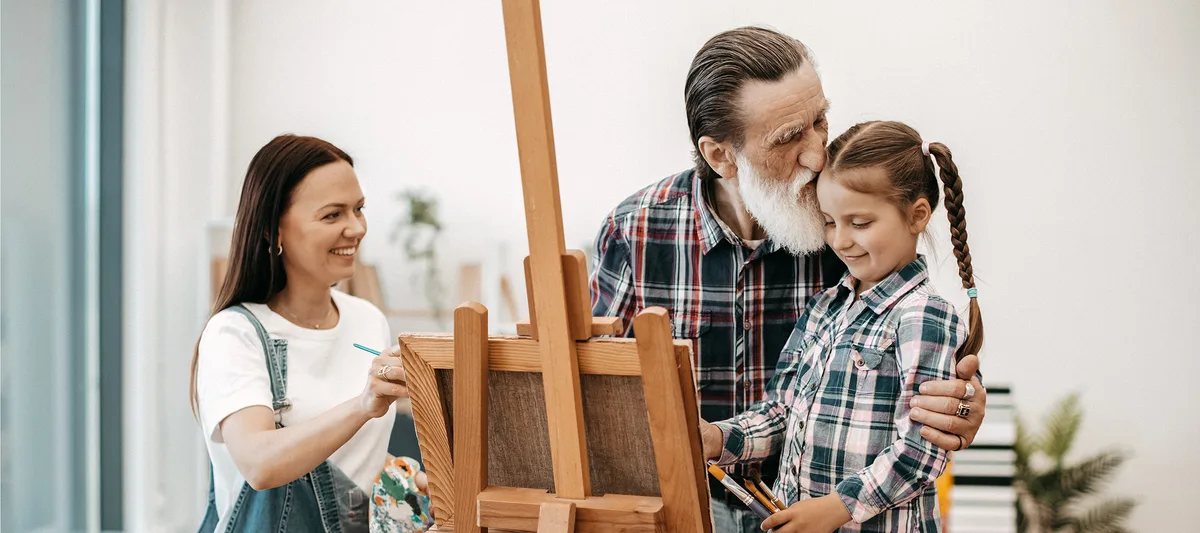 bold-kinder-loben-schweizer-elternmagazin-adobestock-hg Loving aged grandfather in casual wear kissing little kid while smiling lady doing sketches on cloth in room. Affectionate grown-ups and child strengthening family bonds while engaging in arts.