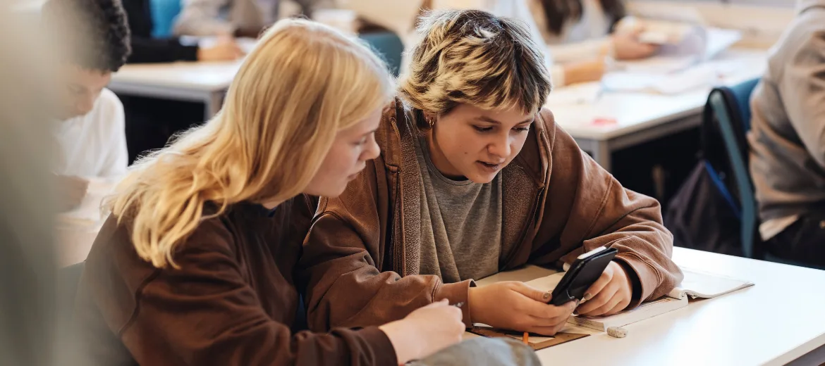 25-03-handy-smartphone-verbot-medienkompetenz-swisscom-schweizer-elternmagazin-fritz-und-fraenzi-hg Zwei Jugendliche sitzen in einem Schluzimmer und schauen auf ein Handy