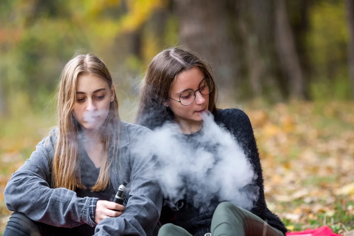 Vape lgbt teenagers. Bisexual lesbian young caucasian teenage girls in casual clothes vape electronic cigarette on the street in the park in the autumn evening. Bad habit. Vapen: Zwei Teenagermädchen sitzen beim Wald auf einer Picknickdecke und vapen.