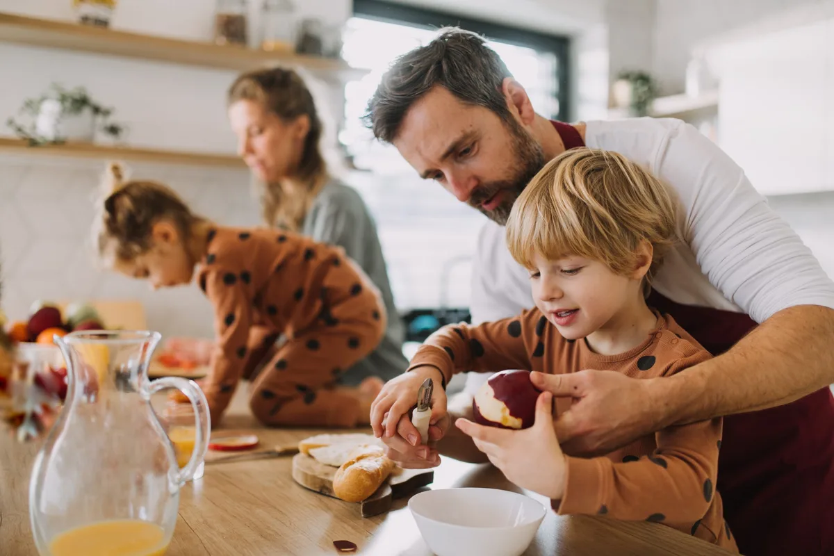 Jeune famille avec deux jeunes enfants préparant ensemble le petit-déjeuner dans la cuisine. Les enfants prennent exemple sur leurs parents lorsqu'ils mangent : père et fils en train de cuisiner