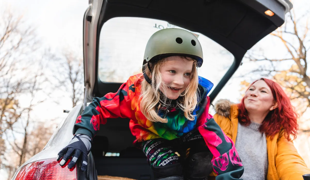 Playful child together with mother sitting in the back of the car. They are doing skateboard outside in a parking road