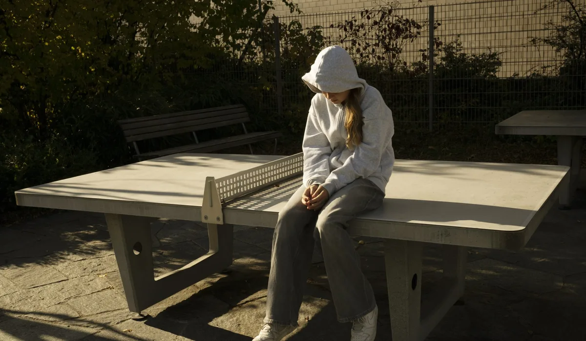 Aggressives Kind, Wut: Teenagermädchen mit Hoodie sitzt auf einem Tischtennistisch aus Beton und blickt frustriert zu Boden.