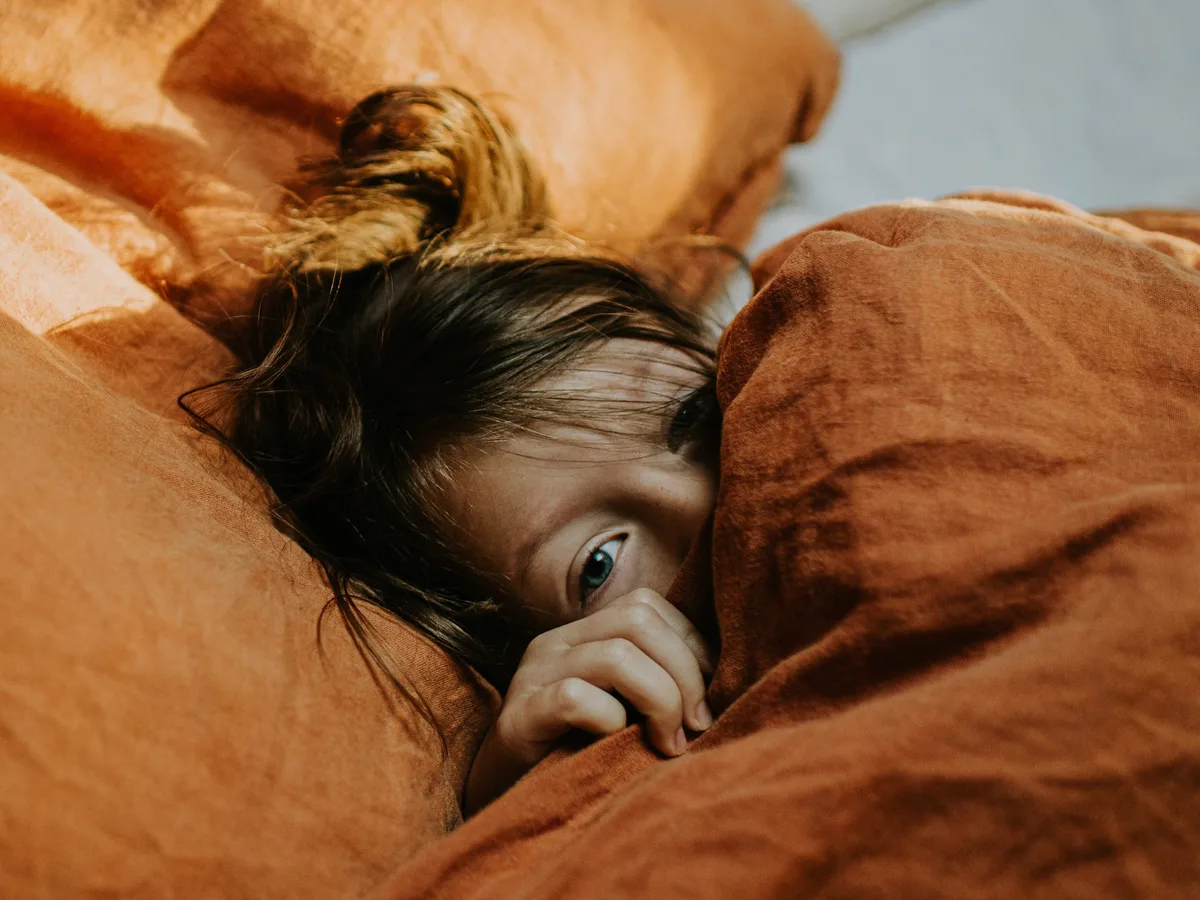 A little girl lies in bed, wrapped in a cozy duvet. She peeks out from under the sheets. A cute young girl lies in a comfy double bed, wrapped in a thick duvet with a terracotta coloured linen duvet cover and white bedding. She looks uncertain as she curiously and tentatively peers out from under the sheets.