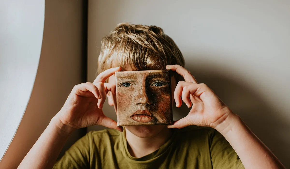 A little boy holds a small oil painting of himself over his face Fazialisparese: Ein blonder Bub hält symbolisch ein gemaltes Gesichtsbild vor sein Gesicht