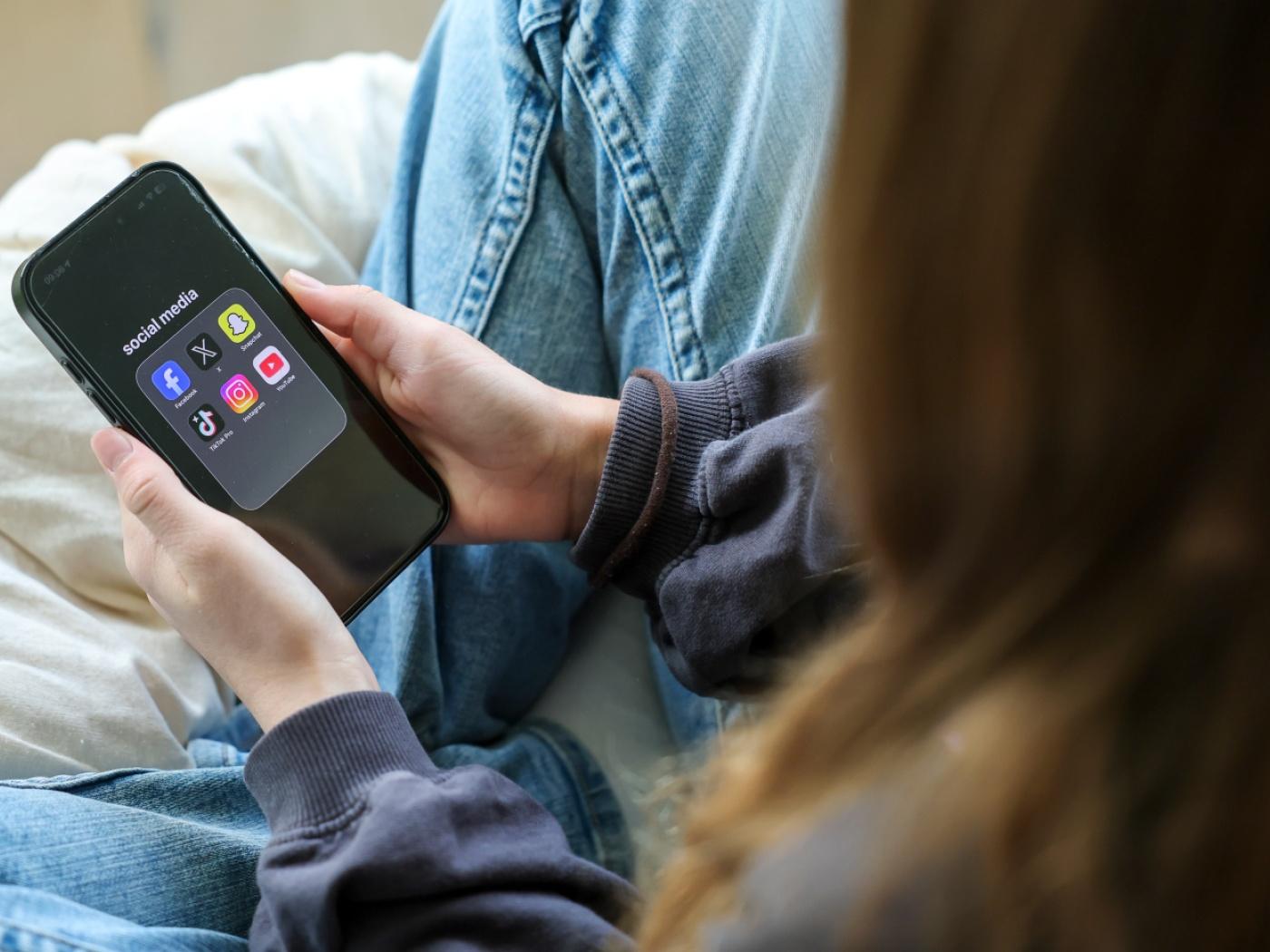 ILLUSTRATION - Ein Mädchen hält ein iPhone 16 in den Händen, auf dessen Display in einem Ordner mit der Bezeichnung "social media" die Logos von Facebook, X (ehemals Twitter), Snapchat, TikTok Pro, Instagram und YouTube zu sehen sind. Foto: Jan Woitas/dpa