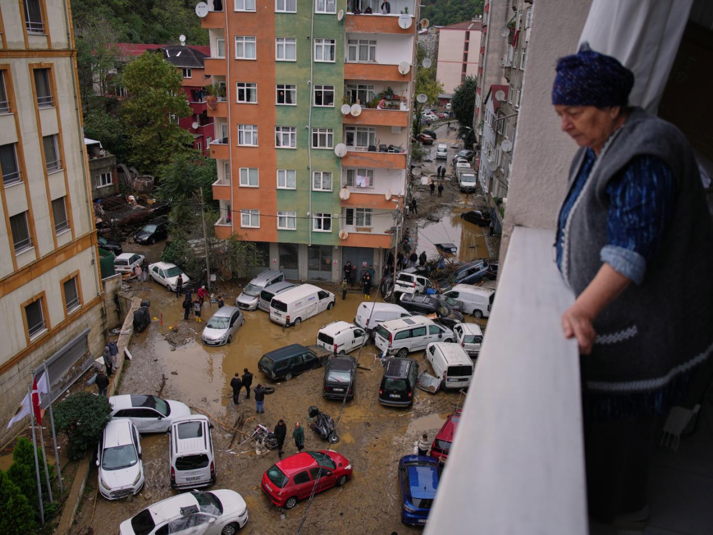 Eine Frau schaut von ihrem Balkon auf Anwohner, die zwischen umgestürzten Fahrzeugen und verstreuten Trümmern umher gehen, nachdem heftige Regenfälle zu Überschwemmungen geführt haben. Foto: Emrah Gurel/AP/dpa