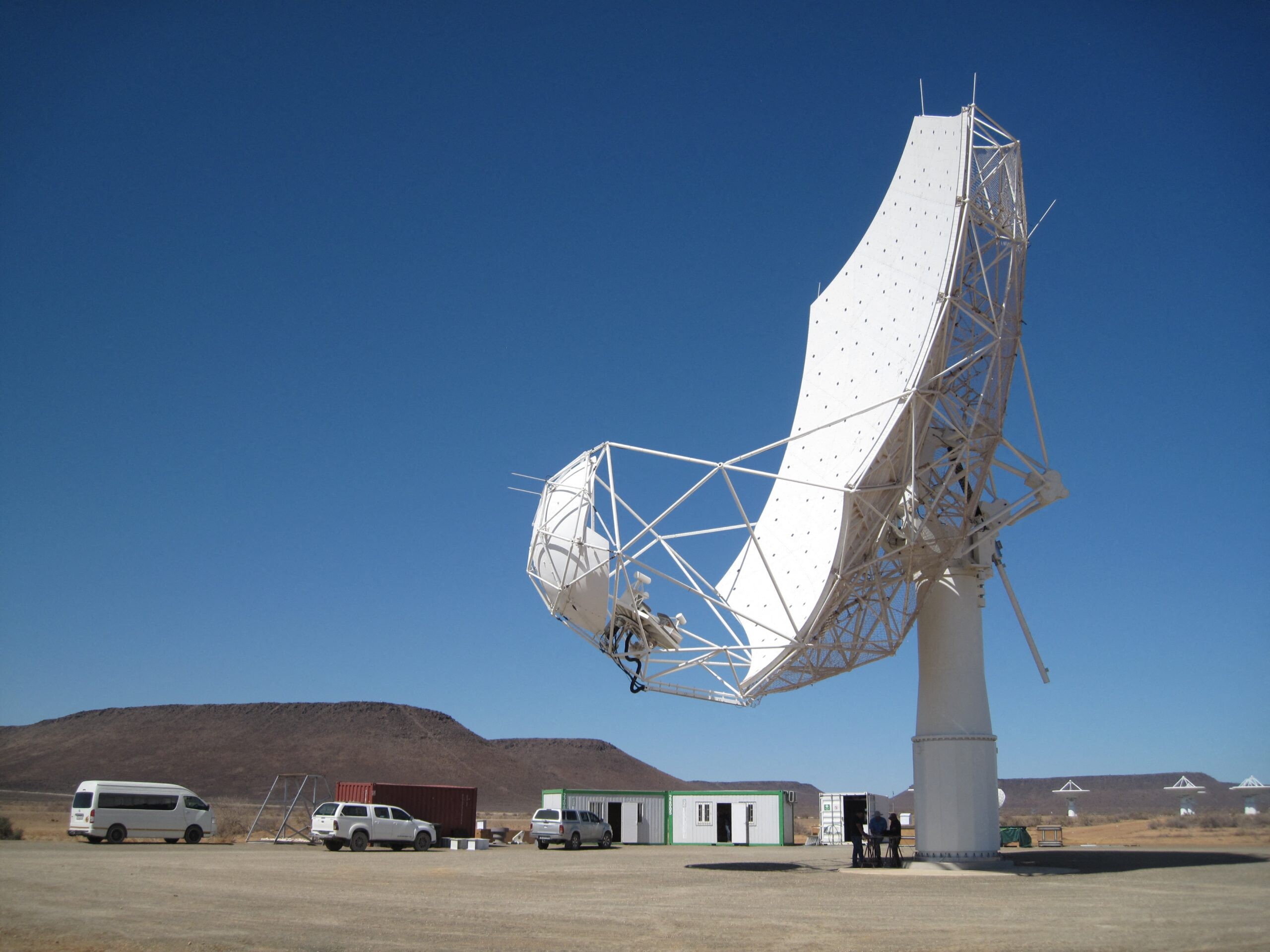 Weiße Radioantenne am SKA-Standort in Südafrika, daneben Fahrzeuge, Container und Personen; weitere Antennen vor kahlen Hügeln im Hintergrund.