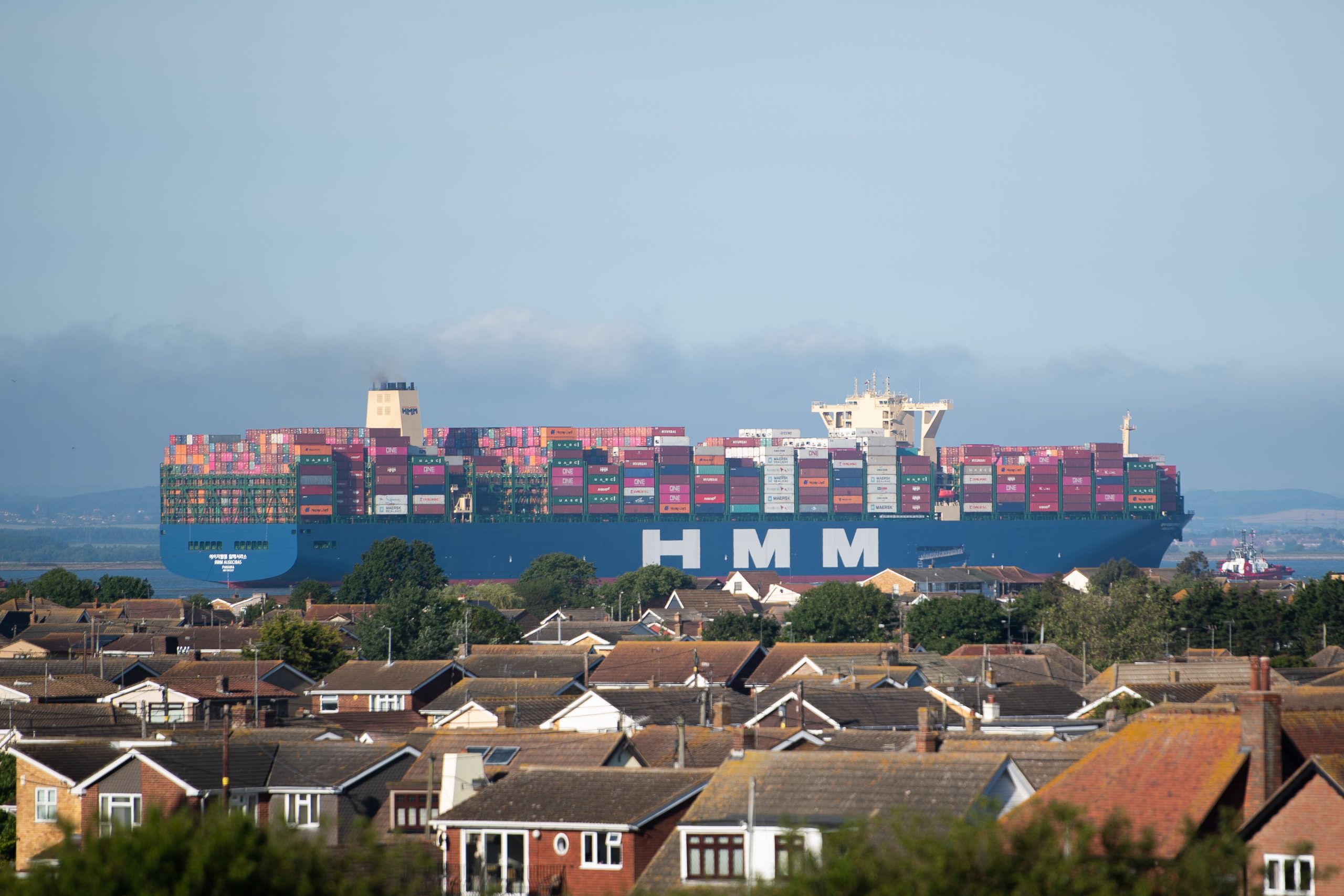 Riesiges Containerschiff HMM Algeciras mit blauem Rumpf und zahlreichen bunten Containerstapeln auf dem Wasser. Im Vordergrund stehen Wohnhäuser, rechts begleitet ein Schlepper das Schiff.