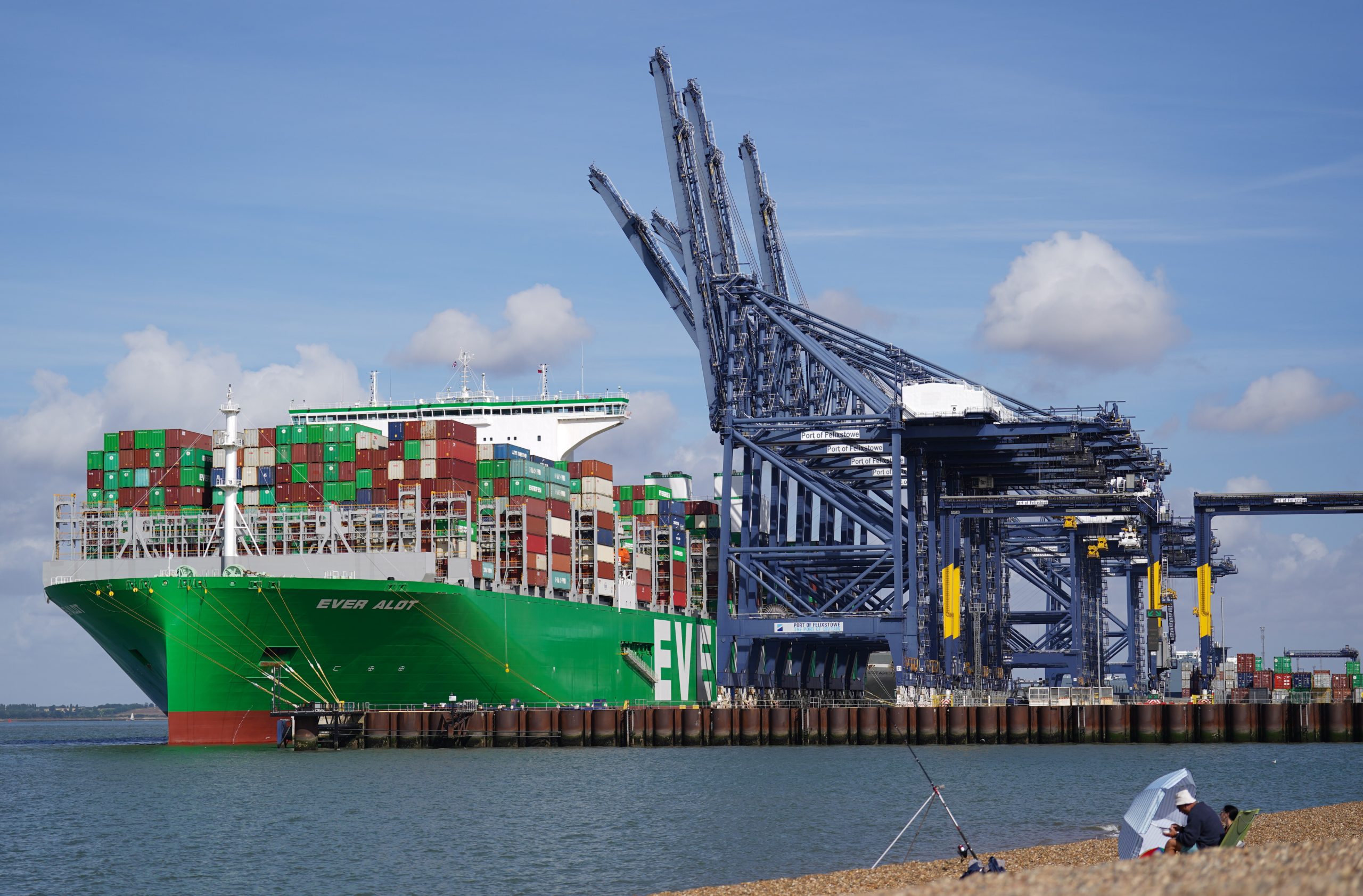 Grünes Containerschiff Ever Alot mit hoch gestapelten Containern am Hafen Felixstowe. Neben dem Schiff stehen große blaue Containerbrücken, im Vordergrund ist ein steiniger Strand zu sehen.