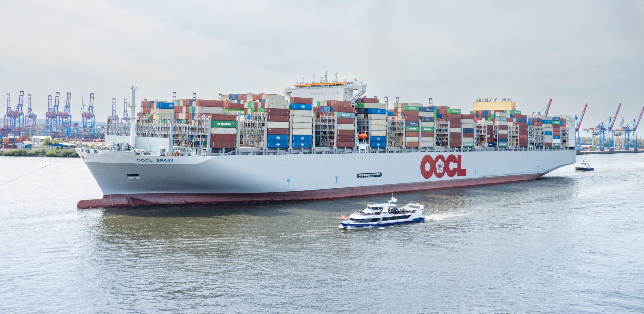 Containerschiff OOCL Spain mit weißem Rumpf und rotem OOCL-Schriftzug in einem Hafen. Zahlreiche bunte Container sind an Bord gestapelt, im Vordergrund fährt ein kleines Passagierschiff.