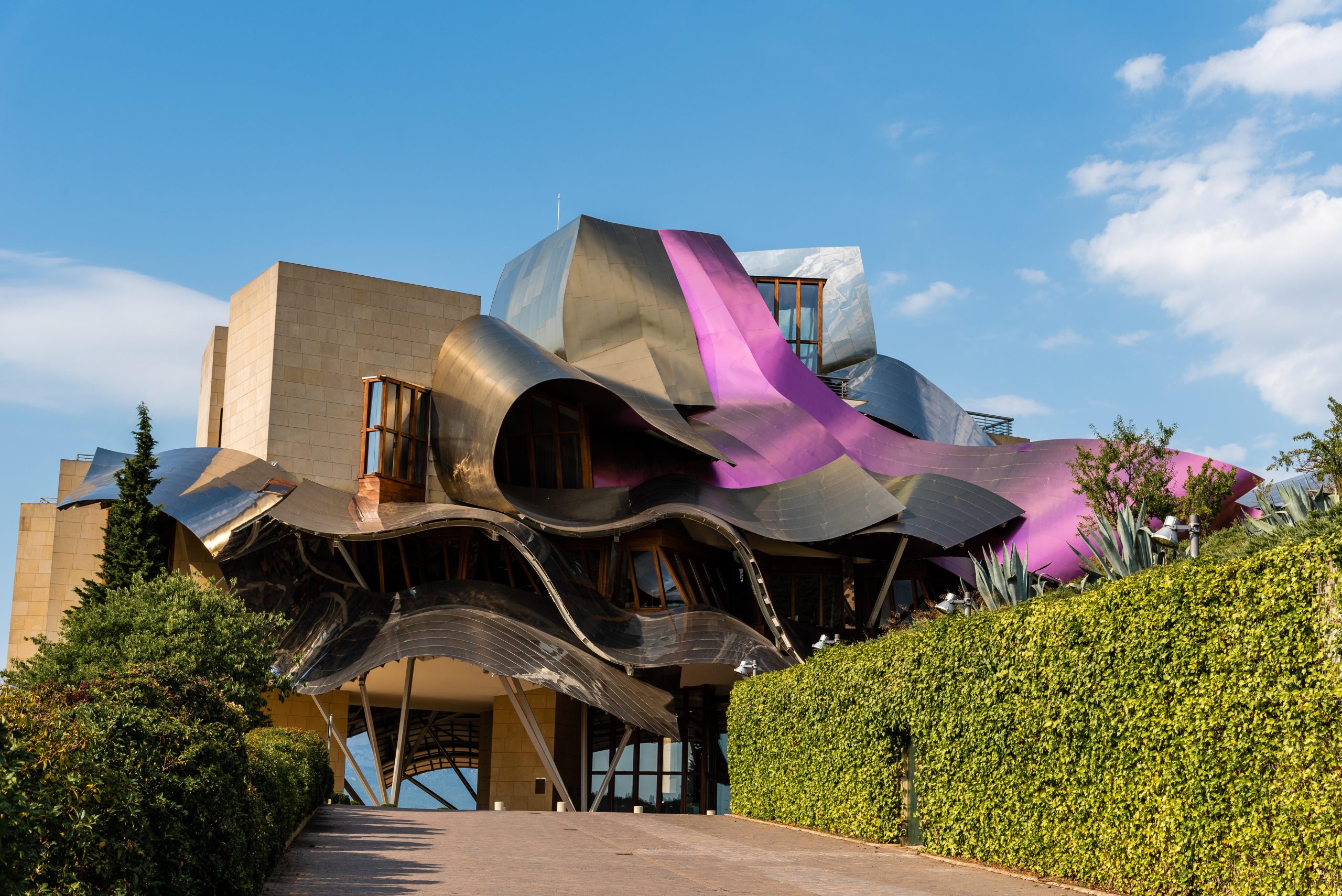 Hotel Marqués de Riscal in Elciego, Spanien, mit geschwungenen Metallfassaden und auffälligen rosafarbenen Titanbändern nach einem Entwurf von Frank Gehry. Im Vordergrund führen ein Weg und grüne Hecken zum Gebäude.