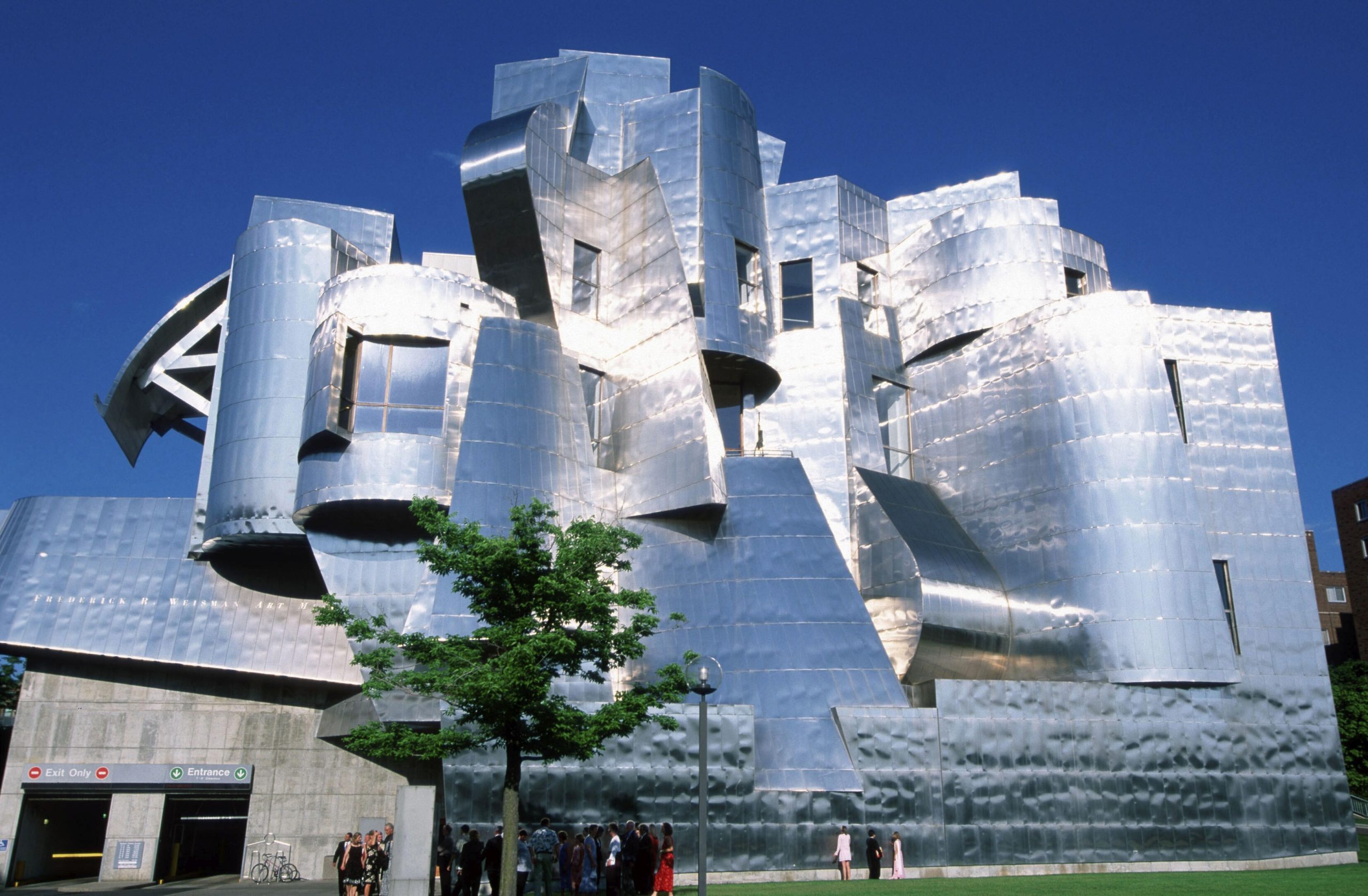 Weisman Art Museum in Minneapolis von Frank Gehry mit silbrig glänzender Edelstahlfassade, asymmetrischen Türmen und geschwungenen Bauteilen. Vor dem Museum stehen ein Baum und mehrere Besucher.