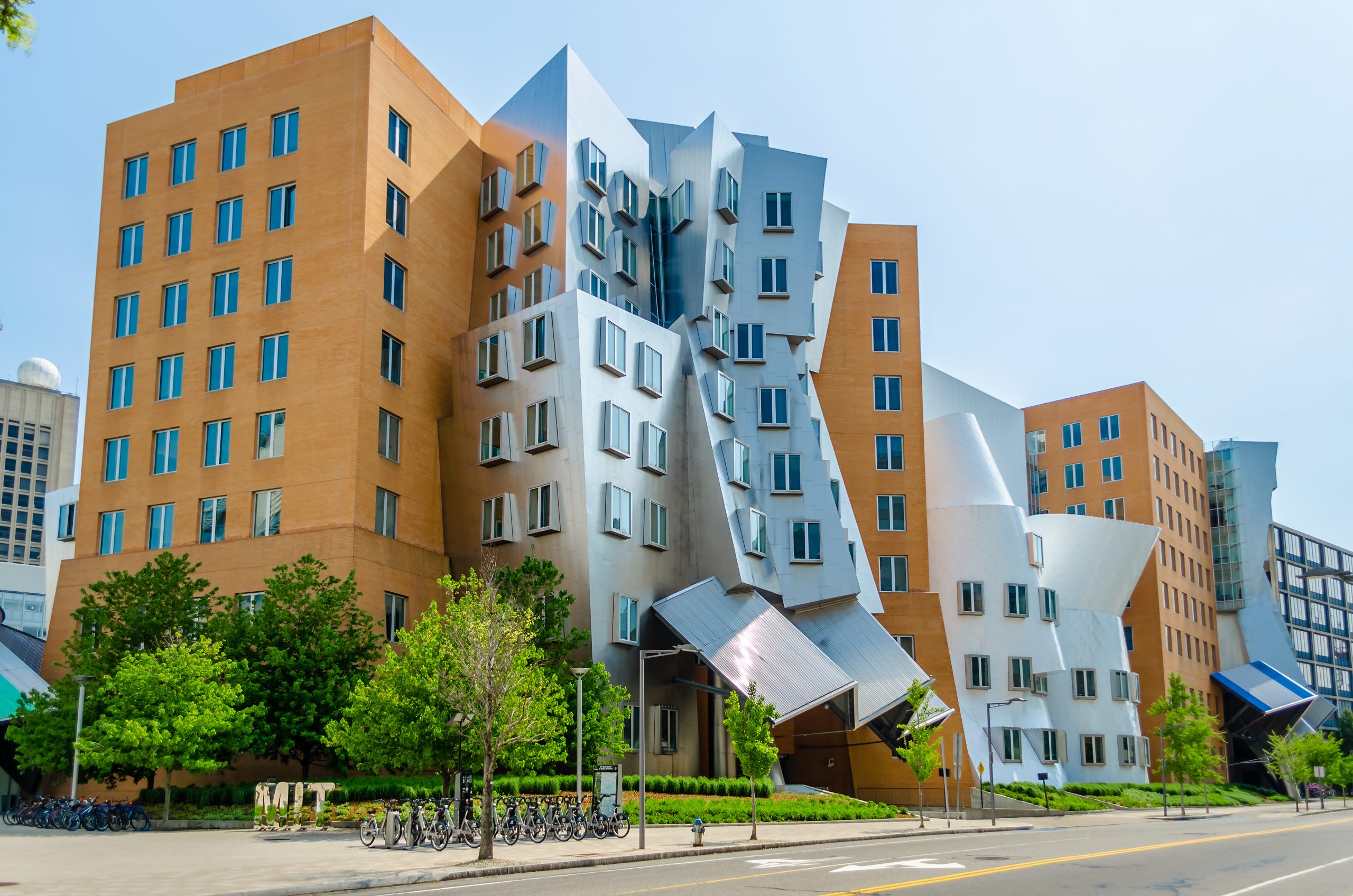 Stata Center am MIT in Cambridge mit schräg angeordneten silbernen Fassaden, hervorstehenden Fenstern und orangefarbenen Gebäudeteilen. Vor dem von Frank Gehry entworfenen Forschungsgebäude stehen Bäume und Fahrräder.