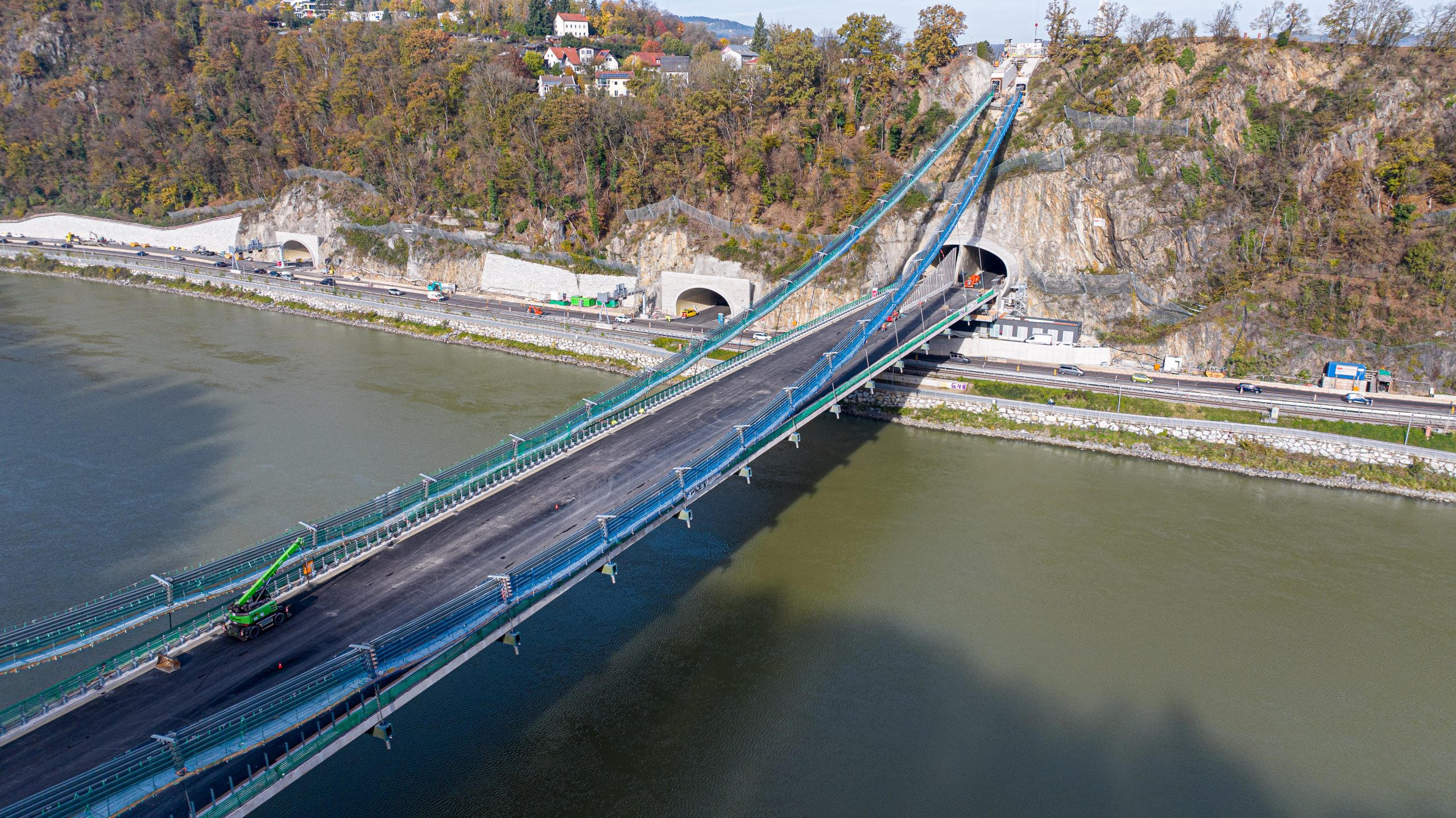 Schräg aufgenommene Luftaufnahme der Donautalbrücke in Linz während der Bauphase. Die breite Fahrbahn hängt an Stahlseilen, die direkt in den Felshang führen. Ein grüner Baukran und Schutzgerüste sind auf der Brücke zu erkennen.