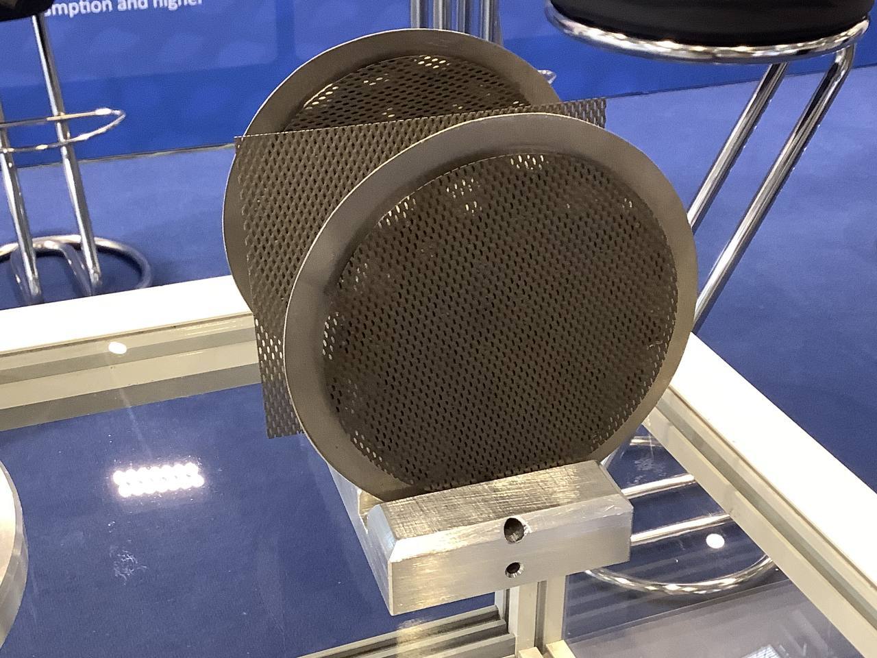 Zwei runde, dunkel beschichtete Elektroden mit gelochter Struktur und ein rechteckiges Stück Streckmetall auf einem Metallhalter in einer Glasvitrine am Messestand.