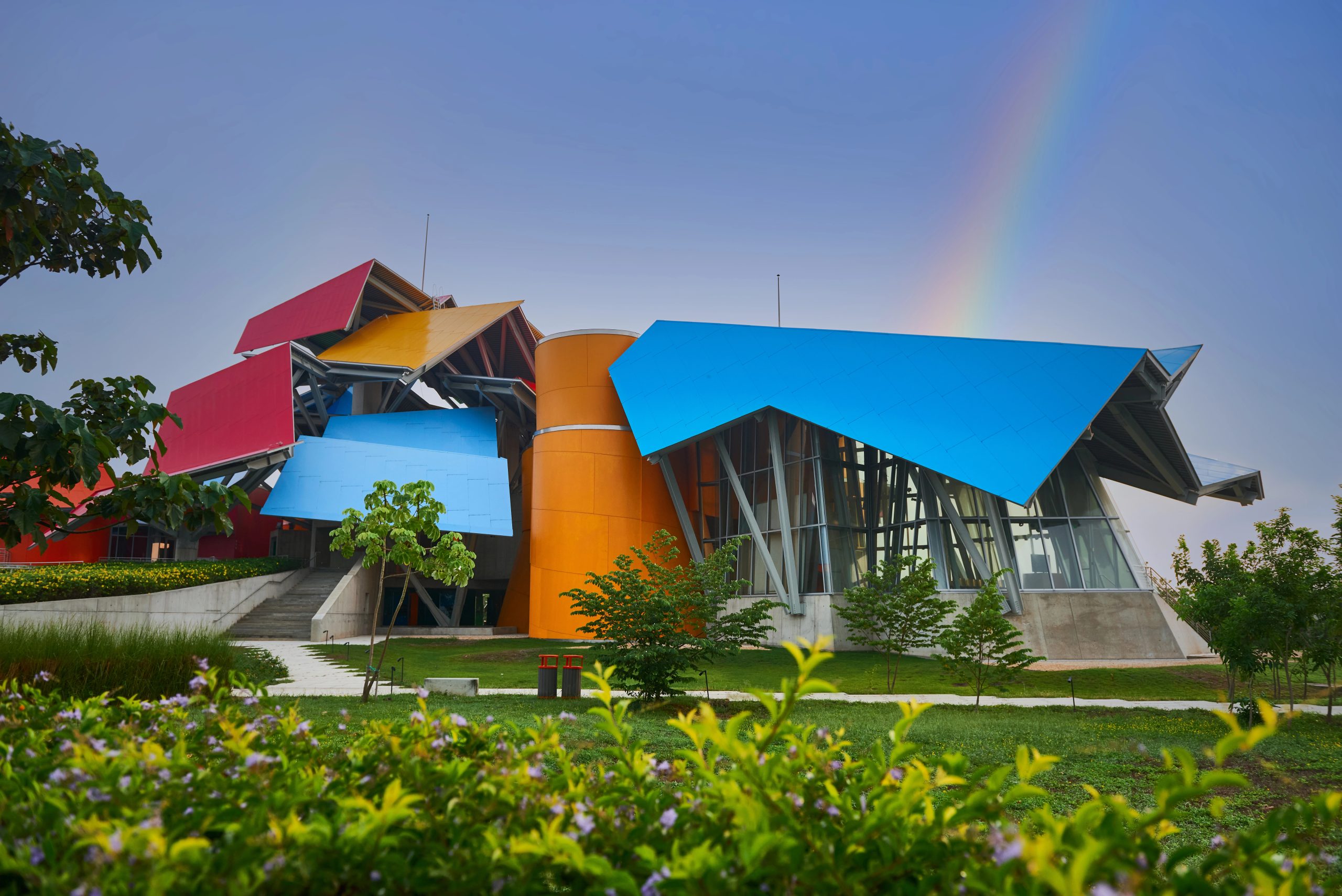 Biomuseo von Frank Gehry in Panama-Stadt mit auffälligen roten, gelben, blauen und orangefarbenen Dachflächen. Vor dem Gebäude liegen Grünflächen und Bäume, am Himmel ist ein schwacher Regenbogen zu sehen.